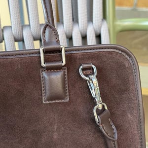 May include: Close-up of a brown suede handbag with a leather handle and silver-tone hardware. The bag features a rectangular shape with a top handle and a detachable shoulder strap. The stitching is visible along the edges.