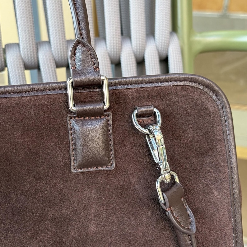 May include: Close-up of a brown suede handbag with a leather handle and silver-tone hardware. The bag features a rectangular shape with a top handle and a detachable shoulder strap. The stitching is visible along the edges.