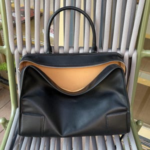 May include: Black leather tote bag with a top handle and a zippered opening. The bag has a rectangular shape with a light brown interior. The bag is sitting on a chair with gray and green accents.