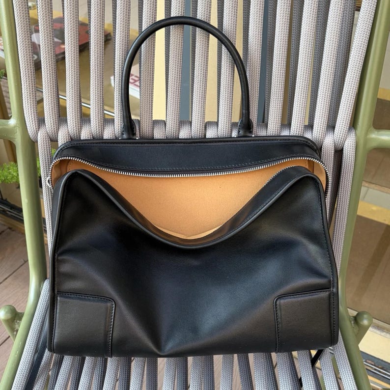 May include: Black leather tote bag with a top handle and a zippered opening. The bag has a rectangular shape with a light brown interior. The bag is sitting on a chair with gray and green accents.