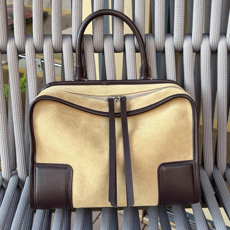 May include: A tan suede handbag with dark brown leather trim and handles. The rectangular bag has a zipper closure and two long, dark brown leather pulls. The bag's corners are reinforced with dark brown leather.