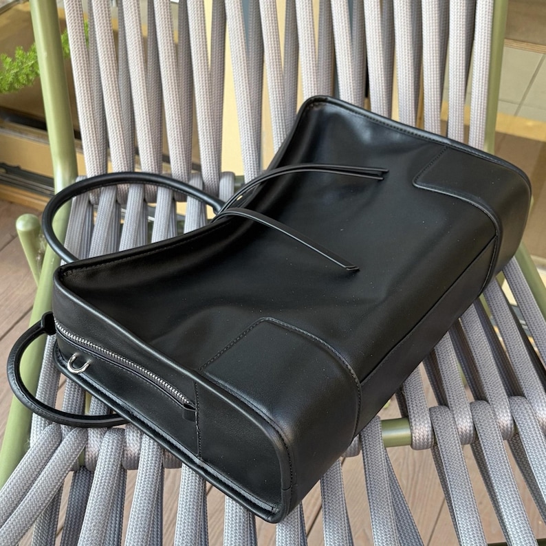 May include: A black leather tote bag with a rectangular shape and a zipper closure. The bag has a handle and a strap. The bag is sitting on a gray chair.
