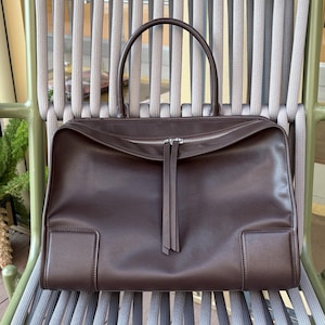 May include: A brown leather handbag with a rectangular shape and a top handle. The bag features a silver zipper closure and two decorative leather tassels. The bag is sitting on a gray and green chair.