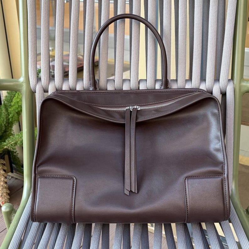 May include: A brown leather handbag with a rectangular shape and a top handle. The bag features a silver zipper closure and two decorative leather tassels. The bag is sitting on a gray and green chair.