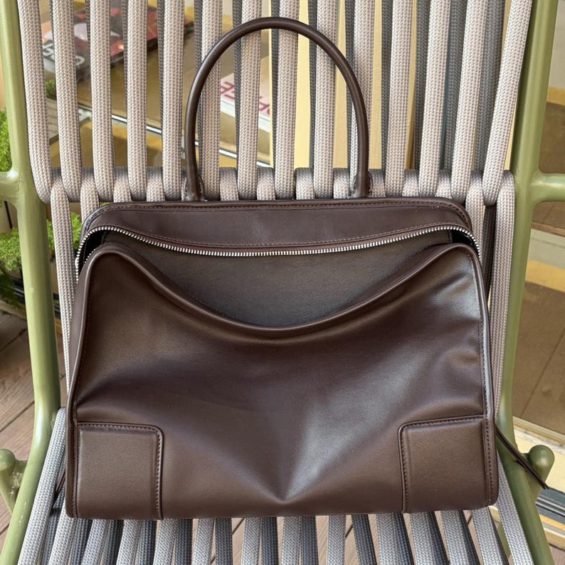 May include: A brown leather handbag with a top handle and a zipper closure. The bag has a rectangular shape with a structured design and is sitting on a chair with a woven back.