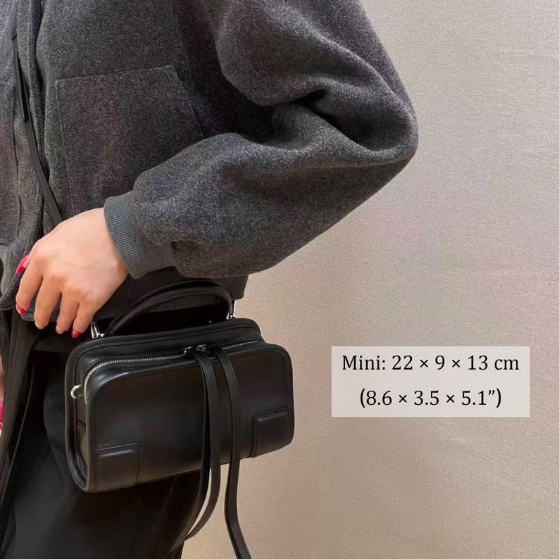 May include: A black mini handbag with a top handle and a long shoulder strap. The bag is rectangular, with a zipper closure and decorative straps. Dimensions are 8.6 x 3.5 x 5.1 inches.