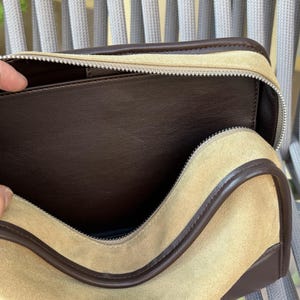 May include: A beige suede travel bag with a brown leather trim and a silver zipper. The interior is a dark brown leather. The bag is open, revealing the inside.