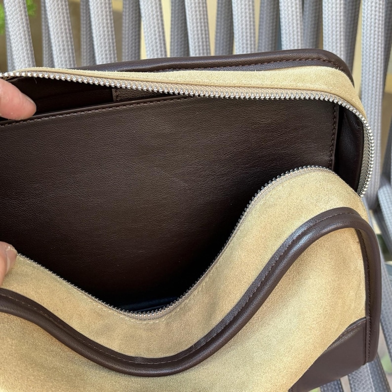 May include: A beige suede travel bag with a brown leather trim and a silver zipper. The interior is a dark brown leather. The bag is open, revealing the inside.