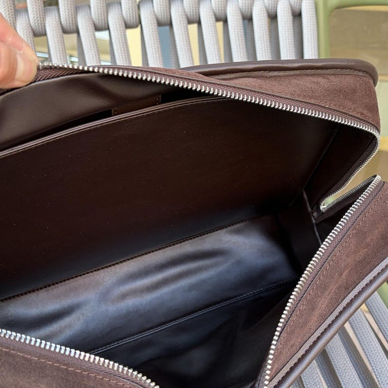 May include: Open view of a brown suede briefcase with a silver zipper. The interior is lined with a darker brown material. The briefcase is open, revealing the inside compartments.