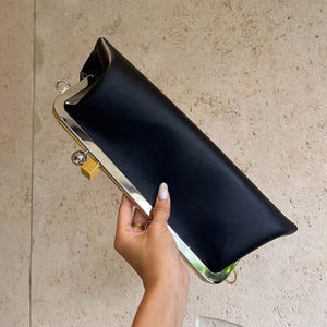May include: A black satin clutch with a silver and gold-tone metal frame. The rectangular bag has a clasp closure and a smooth, elegant appearance. The clutch is held by a hand.