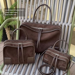 May include: Three brown suede handbags of varying sizes. Each bag has a zipper closure, a top handle, and a leather trim. The smallest bag includes a long, detachable strap. The bags are displayed on a chair.