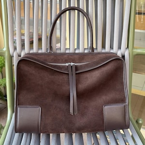 May include: A brown suede handbag with a rectangular shape and a top handle. The bag features a zipper closure and two leather accents on the front. The bag is sitting on a chair with a gray and green design.