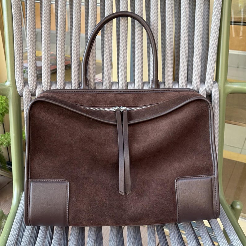 May include: A brown suede handbag with a rectangular shape and a top handle. The bag features a zipper closure and two leather accents on the front. The bag is sitting on a chair with a gray and green design.
