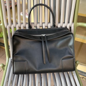 May include: A black leather handbag with a rectangular shape and a top handle. The bag features a silver zipper closure and two long leather zipper pulls. The bag is sitting on a gray and green chair.