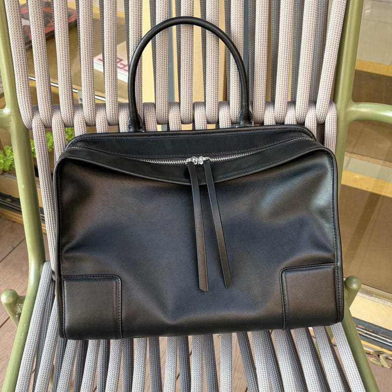 May include: A black leather handbag with a rectangular shape and a top handle. The bag features a silver zipper closure and two long leather zipper pulls. The bag is sitting on a gray and green chair.