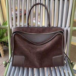 May include: A brown suede handbag with a top handle and a zipper closure. The bag has a rectangular shape with a structured design and a dark brown leather trim. The bag is sitting on a gray and green chair.
