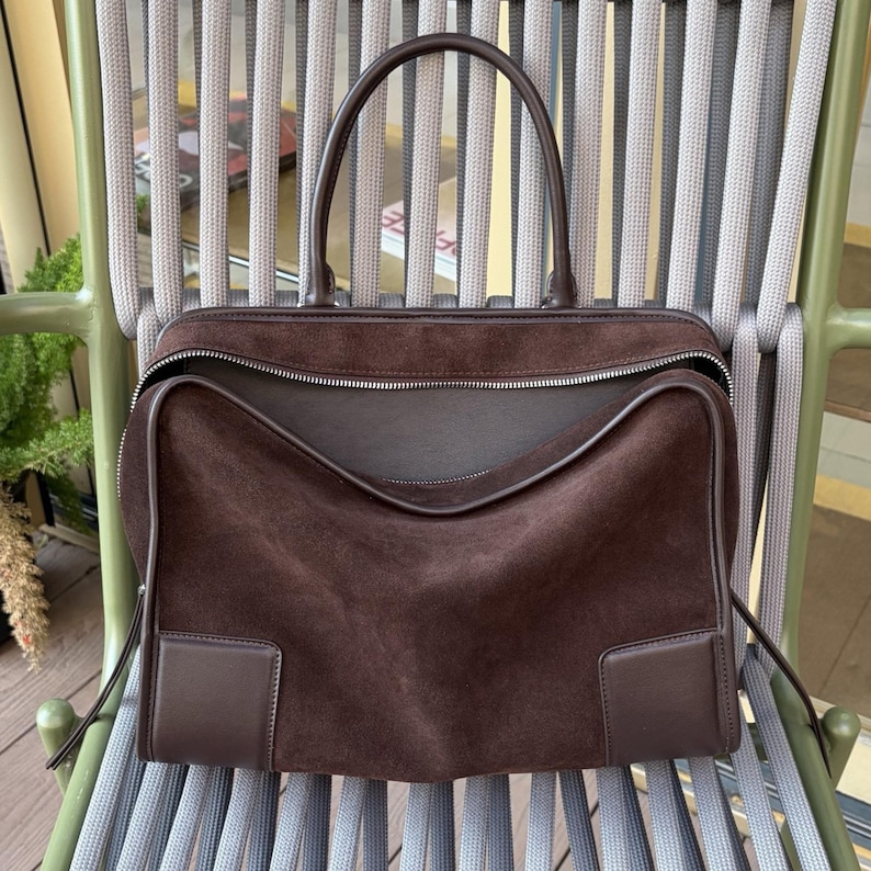 May include: A brown suede handbag with a top handle and a zipper closure. The bag has a rectangular shape with a structured design and a dark brown leather trim. The bag is sitting on a gray and green chair.