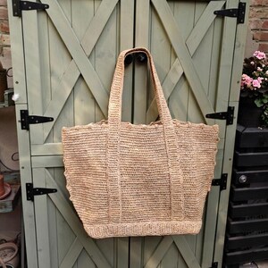 May include: A large, woven straw tote bag with strong handles. The bag has a rectangular shape with a scalloped edge, hanging against a light green wooden door. The natural straw colour contrasts with the dark hinges.
