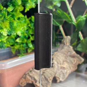 May include: A black, vertically oriented handle with a textured, ribbed surface attached to a clear glass panel. The handle features a concave grip area running down its centre, providing a functional grasp for sliding the glass door open or closed.