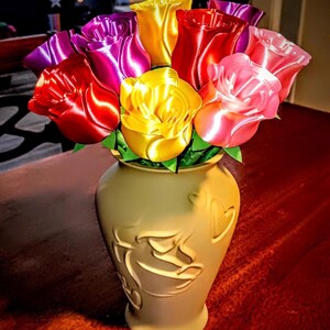 Puede incluir: Un ramo de rosas de cinta coloridas en un jarr&oacute;n color crema. Las rosas son en tonos de rojo, rosa, amarillo y morado. El jarr&oacute;n tiene un dise&ntilde;o decorativo en relieve. El arreglo est&aacute; sobre una superficie roja oscura.