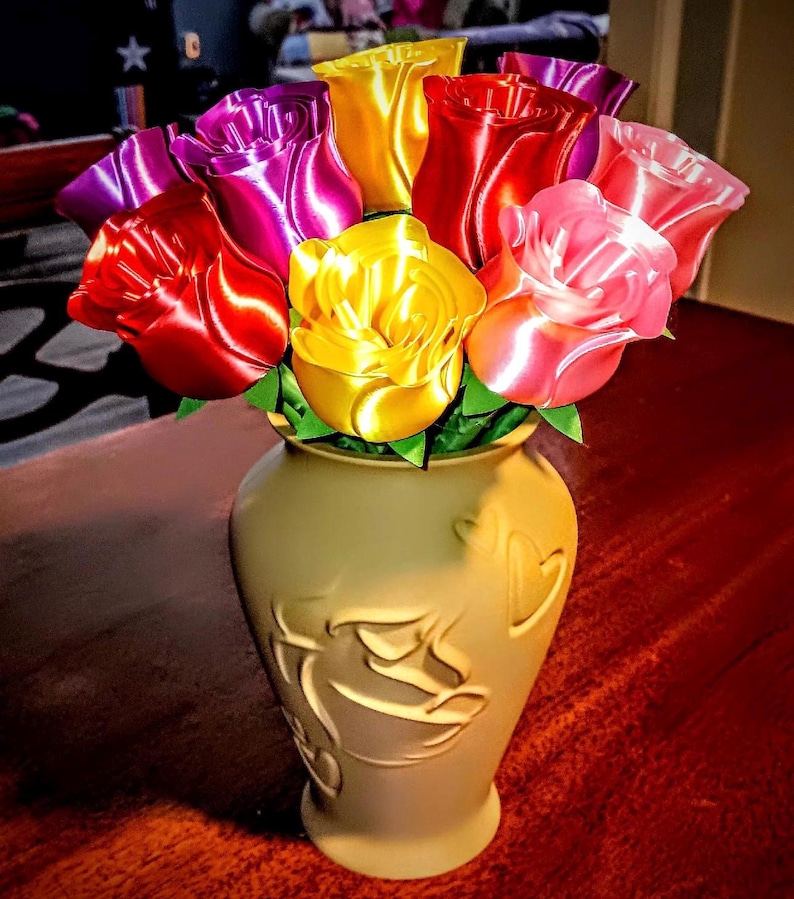 Puede incluir: Un ramo de rosas de cinta coloridas en un jarr&oacute;n color crema. Las rosas son en tonos de rojo, rosa, amarillo y morado. El jarr&oacute;n tiene un dise&ntilde;o decorativo en relieve. El arreglo est&aacute; sobre una superficie roja oscura.
