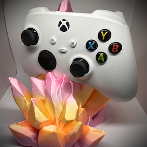 May include: A white Xbox controller is displayed on a pink and yellow crystal-shaped stand. The controller has the standard Xbox buttons, including A, B, X, and Y.
