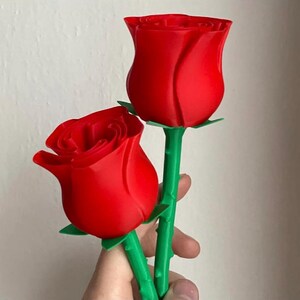 Puede incluir: Dos capullos de rosas rojas con tallos y hojas verdes. Las rosas son de un color rojo vibrante y los tallos tienen una apariencia realista. Las rosas son un objeto decorativo hermoso y elegante.