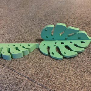 May include: Two green monstera leaf-shaped wall decor pieces. The larger piece is a full leaf with a hole in the center. The smaller piece is half a leaf.