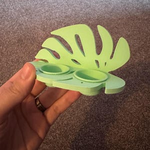 以下が含まれることがあります： モンスターの葉の形をした、薄い緑色の3Dプリントされたプランター。小さな丸い鉢が2つ付いています。