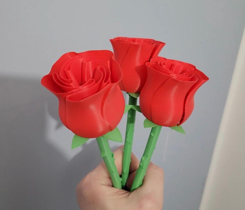 Puede incluir: Tres tallos de rosas rojas con tallos y hojas verdes. Las rosas son de un color rojo vibrante, con p&eacute;talos detallados. Los tallos se sostienen en una mano, sobre un fondo gris. Las rosas parecen estar impresas en 3D.