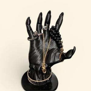 May include: A black hand-shaped jewellery display stand featuring long, pointed fingers. The stand is adorned with a gold-tone chain necklace, a black beaded bracelet, two rings on the fingers, and two stacked bracelets around the wrist base.