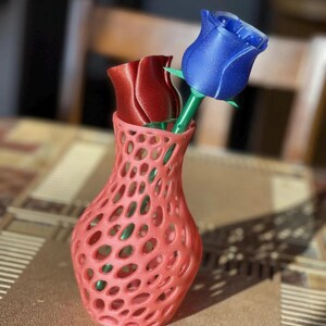 Puede incluir: Un jarr&oacute;n calado de color coral contiene dos rosas artificiales. Una rosa es de color rojo intenso, la otra azul brillante. Los tallos son verdes. El jarr&oacute;n est&aacute; sobre una mesa con una superficie estampada.
