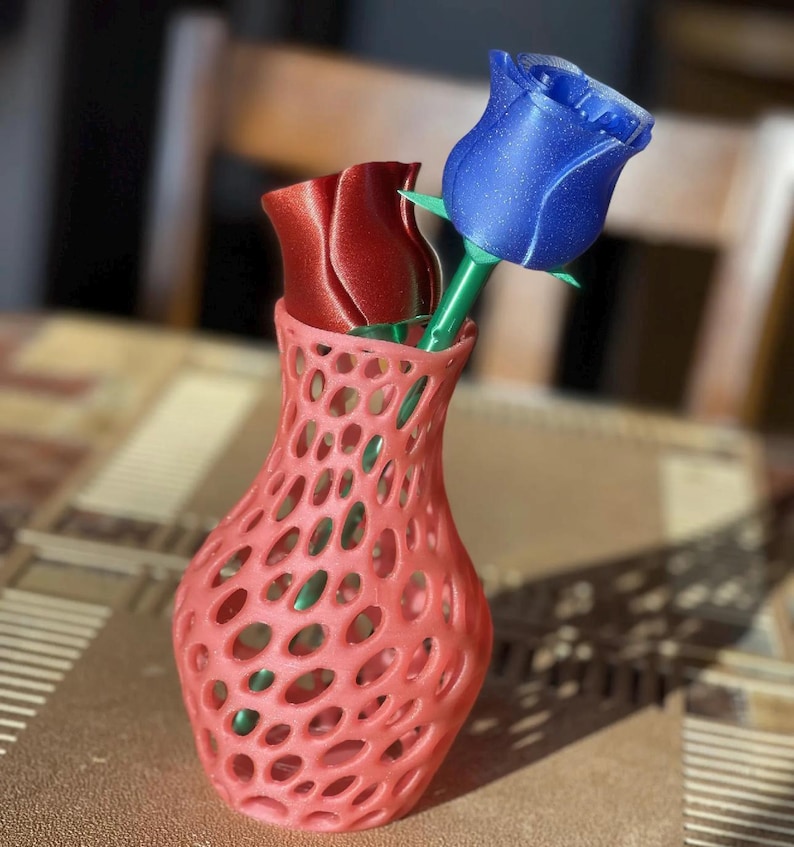 Puede incluir: Un jarr&oacute;n calado de color coral contiene dos rosas artificiales. Una rosa es de color rojo intenso, la otra azul brillante. Los tallos son verdes. El jarr&oacute;n est&aacute; sobre una mesa con una superficie estampada.