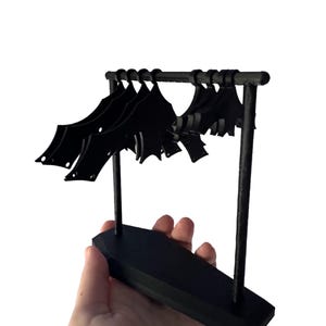 Peut inclure: Un support en plastique noir avec une rangée de cintres en forme de chauve-souris noirs. Le support a une base rectangulaire et deux supports verticaux.