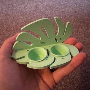 以下が含まれることがあります： 薄い緑色の3Dプリントされたモンステラの葉の形をした皿で、2つの小さな丸い窪みのある区画があります。皿は人の手に持たれています。