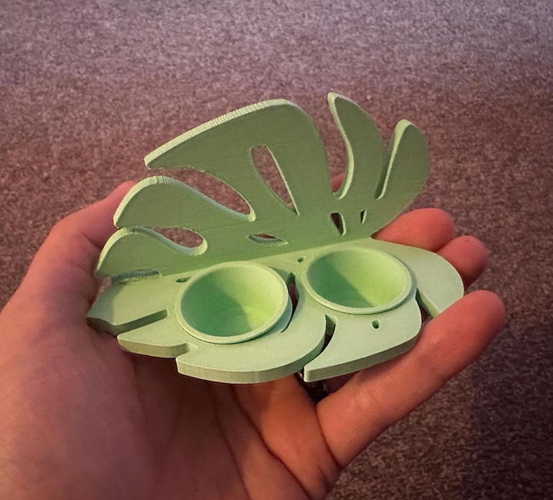 以下が含まれることがあります： 薄い緑色の3Dプリントされたモンステラの葉の形をした皿で、2つの小さな丸い窪みのある区画があります。皿は人の手に持たれています。