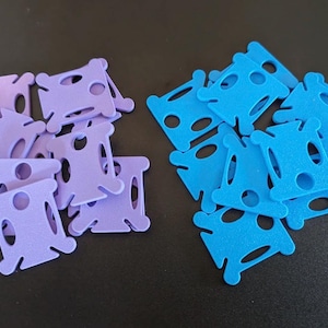 May include: Two groups of thread bobbins. One group is light purple, the other bright blue. Each bobbin has a distinctive shape with holes and slots for holding thread. These are likely used for cross-stitch or embroidery. Measurements are in cm.