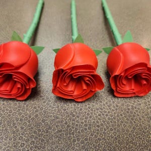Puede incluir: Tres capullos de rosas rojas con tallos y hojas verdes dispuestos en fila. Las rosas tienen p&eacute;talos detallados y est&aacute;n sobre un fondo texturizado de tonos neutros. Los tallos son verdes.