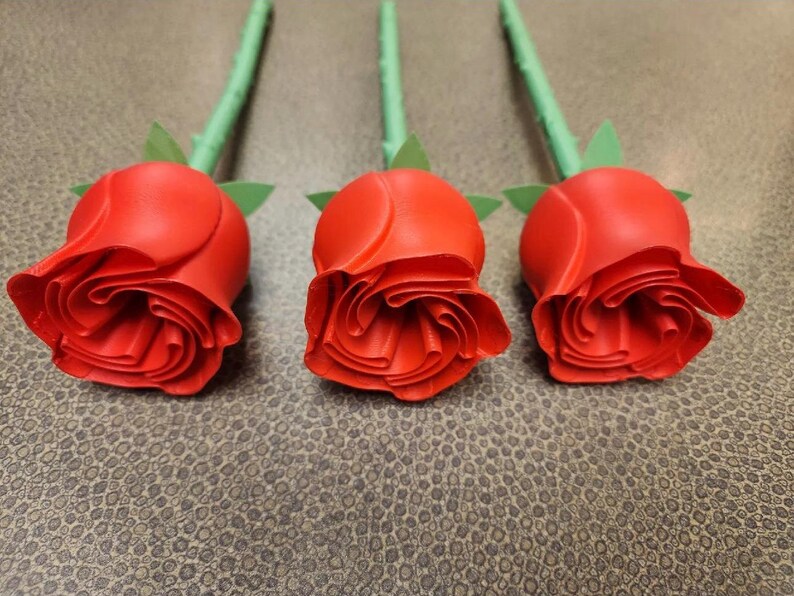 Puede incluir: Tres capullos de rosas rojas con tallos y hojas verdes dispuestos en fila. Las rosas tienen p&eacute;talos detallados y est&aacute;n sobre un fondo texturizado de tonos neutros. Los tallos son verdes.