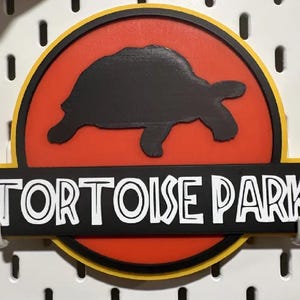 May include: A sign with a black tortoise silhouette on a red circle, with a yellow and black border. The sign reads "TORTOISE PARK" in white letters on a black rectangle. The sign is mounted on a white pegboard.