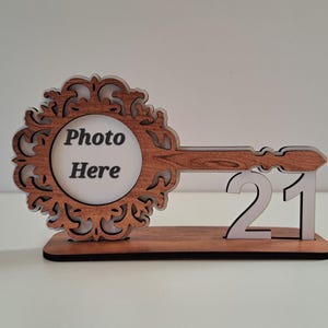 Puede incluir: Un marco de fotos de madera con forma de llave, con el número 21. El marco tiene un diseño ornamental alrededor del área circular de la foto, con las palabras "Photo Here" impresas en el centro. El marco es de madera marrón clara y oscura.
