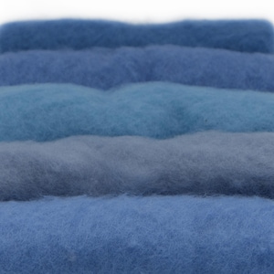 Op de afbeelding: Een verzameling van vijf horizontale stroken ruwe wolvezels gerangschikt in een stapel. De vezels vertonen een verloop van blauwtinten, variërend van diep marineblauw en middenblauw tot lichter hemelsblauw en een gedempte, grijsblauwe tint.