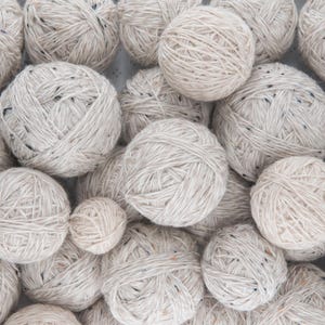 May include: A collection of off-white yarn balls with flecks of color throughout. The yarn is tightly wound, creating a textured surface. The balls vary in size, suggesting a variety of projects. Ideal for knitting or crocheting.