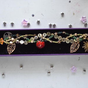 May include: A multi-strand bracelet featuring green and gold beads, star-shaped charms, two leaf charms, a sun charm, and a ladybug charm. The bracelet includes letter beads spelling out the phrase THE GREAT DIVIDE in a purple gift box.