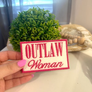May include: A rectangular patch with the words "OUTLAW Woman" embroidered in pink on a white background. The patch has a pink border. A small green plant is in the background.