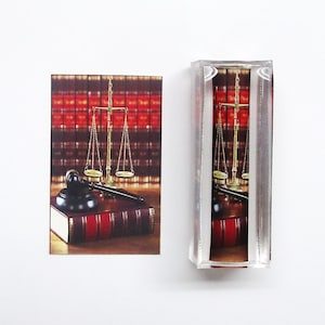 May include: A gold scale of justice with a gavel and a stack of law books in front of a red and black bookshelf.