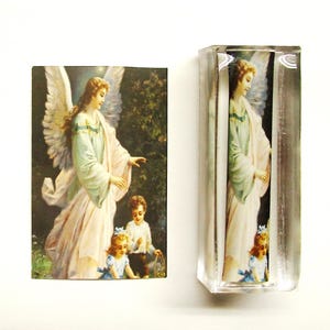 May include: A glass block with a printed image of an angel with wings, wearing a white gown, and two children. The angel is holding out her hand towards the children. The image is set against a green background with trees.