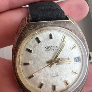 Op de afbeelding: Vintage Gruen Precision horloge met een zilveren wijzerplaat, gouden wijzers en rechthoekige uurmarkeringen. Het horloge heeft een datumweergave en is gemarkeerd met "17 Jewels Autowind". Het heeft een zwarte leren band en een aangetaste metalen kast.