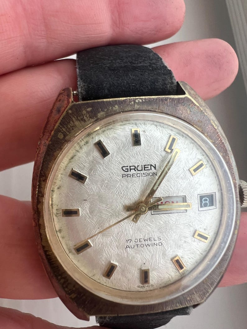 Op de afbeelding: Vintage Gruen Precision horloge met een zilveren wijzerplaat, gouden wijzers en rechthoekige uurmarkeringen. Het horloge heeft een datumweergave en is gemarkeerd met "17 Jewels Autowind". Het heeft een zwarte leren band en een aangetaste metalen kast.