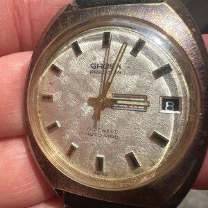 Op de afbeelding: Vintage Gruen Precision polshorloge met een getextureerde zilveren wijzerplaat, goudkleurige wijzers en markeringen, en een datumvenster. Het horloge heeft een zwarte leren band en de tekst "17 JEWELS AUTOWIND" is zichtbaar.